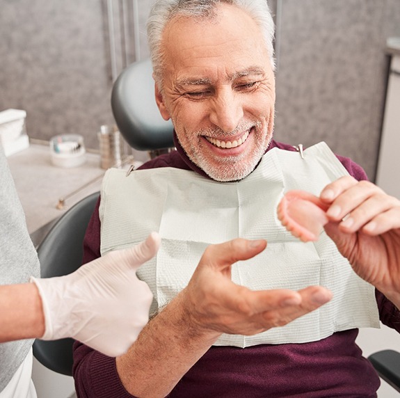 Man in dental chair smiling holding dentures with dentist’s hand giving thumbs up